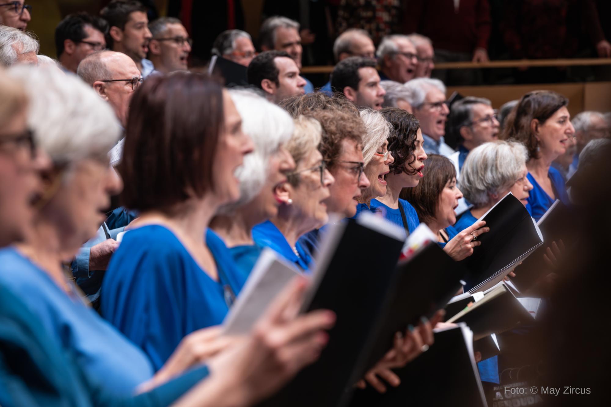 Primer pla lateral d’un cor en concert: diverses cantaires amb roba blava canten mentre sostenen partitures obertes. Al fons, en segon terme, es veuen més membres del cor (també homes) cantant en grades, amb el públic i la sala desenfocats. A la cantonada inferior dreta hi consta el crèdit “Foto: © May Zircus”.