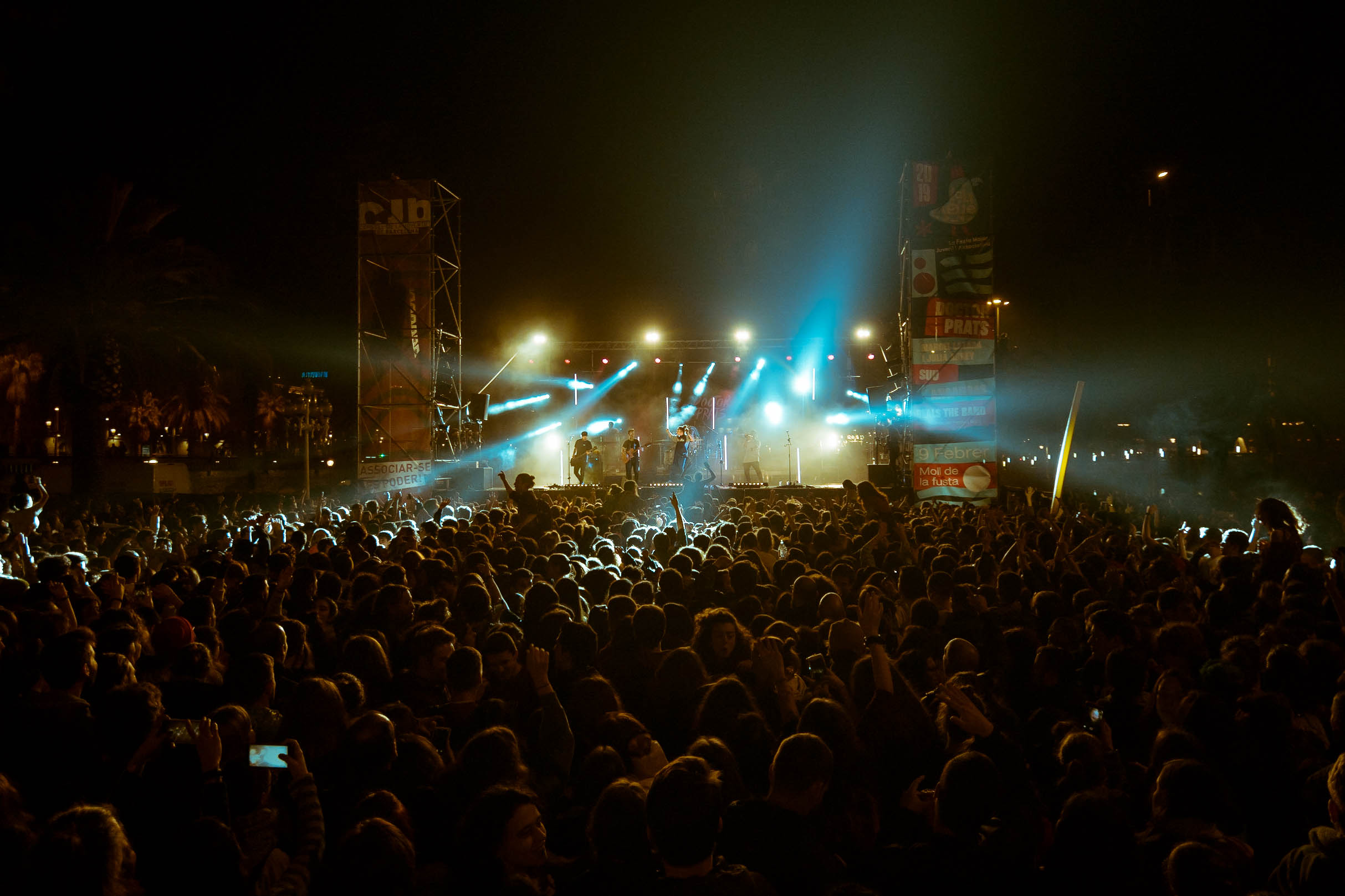 Una gran multitud de persones gaudint d'un concert a l'aire lliure de nit. L'escenari està il·luminat amb llums brillants de diversos colors, i es poden veure músics actuant mentre el públic aixeca les mans.