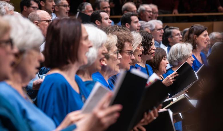 Primer pla lateral d’un cor en concert: diverses cantaires amb roba blava canten mentre sostenen partitures obertes. Al fons, en segon terme, es veuen més membres del cor (també homes) cantant en grades, amb el públic i la sala desenfocats. A la cantonada inferior dreta hi consta el crèdit “Foto: © May Zircus”.