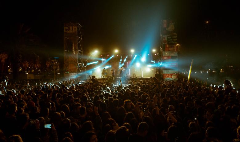 Una gran multitud de persones gaudint d'un concert a l'aire lliure de nit. L'escenari està il·luminat amb llums brillants de diversos colors, i es poden veure músics actuant mentre el públic aixeca les mans.