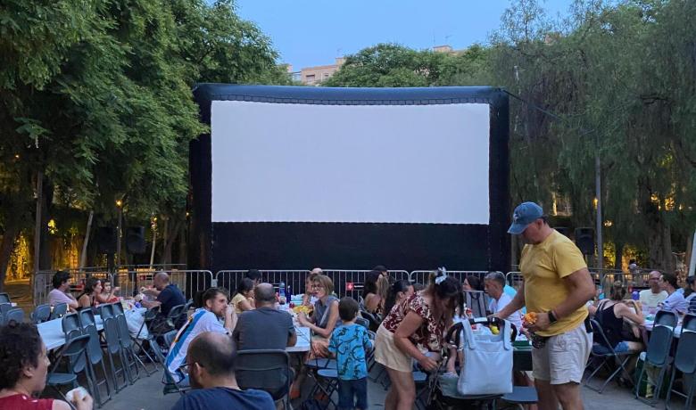 a la imatge es veu una pantalla inflable i gent asseguda en cadires, algunes d'elles sopant