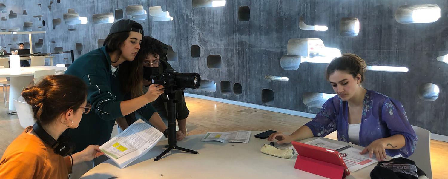 En una sala lluminosa, quatre persones col·laboren al voltant d’una taula durant un procés de gravació audiovisual. Al centre, una càmera sobre un trípode és ajustada per dues persones. La resta de l’equip revisa guions i continguts en paper, coordinant-se per preparar la gravació.